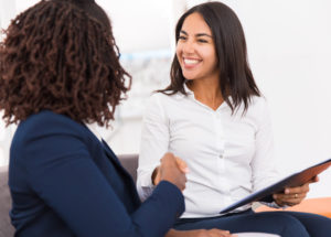 Happy diverse business partners closing deal. Businesswomen sitting on couch, holding folder with documents, shaking hands and smiling. Agreement concept